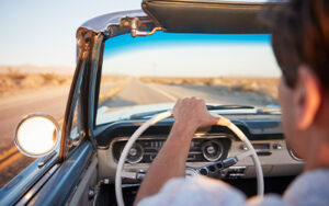 A person drives a classic 1950's car down an open desert road.
