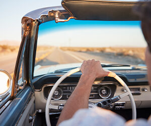 A person drives a classic 1950's car down an open desert road.