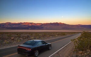 A black Dodge on a wide open road with purple mountains in the distance.