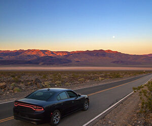 A black Dodge on a wide open road with purple mountains in the distance.
